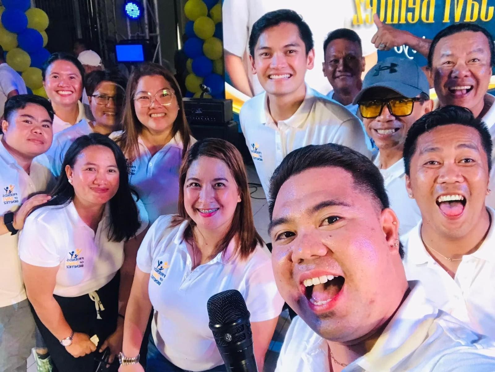 Group of people smiling and posing together, wearing matching white polo shirts at an indoor event.