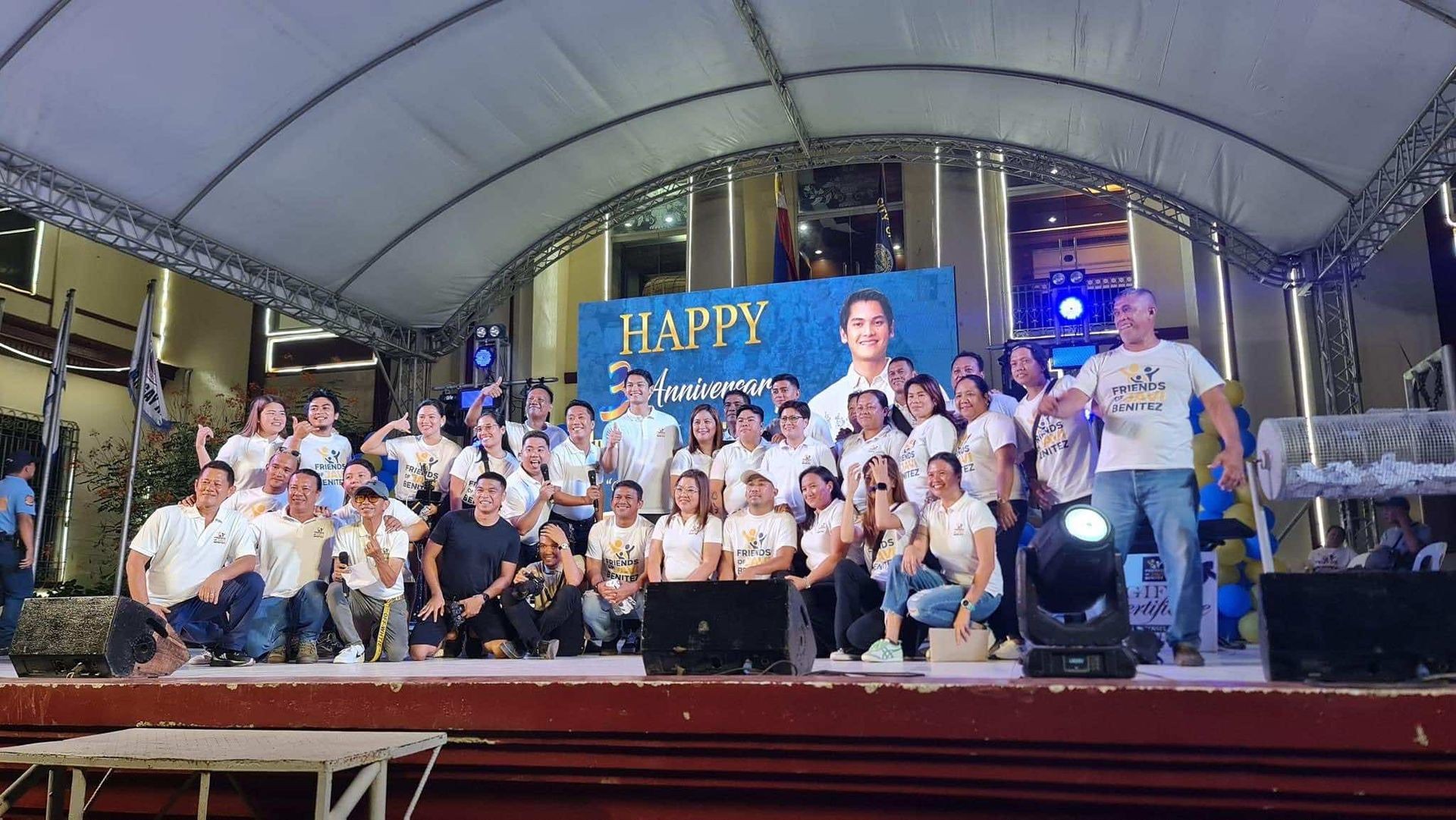 Group of people on stage celebrating an anniversary with a large screen and lights overhead.